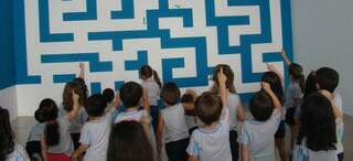 Crianças desvendam labirinto gigante, desenhado na parede.