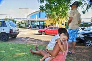 No canteiro em frente à UPA da Vila Almeida, Mário (em pé) foge do calor do posto e encara espera ao lado da mãe e do irmão criança. (Foto: Fernando Antunes)