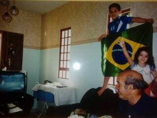 Comemoração do penta na casa de Renan. Ele segura a bandeira do Brasil ao lado da irmã, e seu pai, José, olha atento para a televisão. (Foto: Acervo Pessoal)