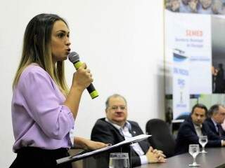 Tatiana Trad discursa em evento público, observada pelo marido, ao fundo. (Foto: Divulgação/PMCG)