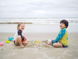 Maria Flor e Vicente brincando na areia da praia, de frente para o mar. (Foto: Arquivo pessoal)