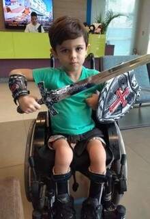 Ele sentado em sua cadeira de rodas e segurando a espada e escudo de herói (Foto: Arquivo pessoal)