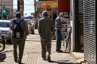 Idoso entrevistado pelo Campo Grande News trabalhando como vendedor em rua do Centro, enquanto outros idosos caminham próximos a ele (Foto: Kísie Ainoã)