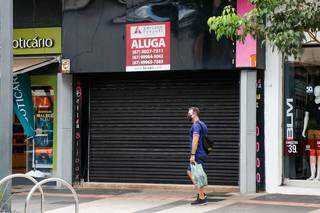 Homem, de máscara, caminha na frente de loja fechada no Centro de Campo Grande (Foto: Henrique Kawaminami)