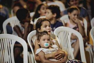 Menina com outra criança no colo assistindo peça de teatro. (Foto: André Patroni)