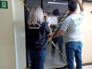 Fiscal do Procon e policial civil lacrando porta da empresa nesta quinta-feira. (Foto: Divulgação | Procon-MS)
