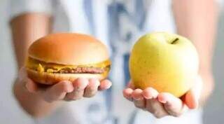 Lanche e fruta lado a lado nas mãos de uma criança, escola que pode evitar obesidade infantil. (Foto: Divulgação/Fiocruz)