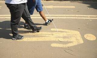 Pessoa cega caminha ao lado de outra pessoa, em local sem piso tátil. (Foto: Agência Brasil/Reprodução)