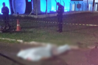 Corpo da vítima coberto por lençol e policial no local do acidente (Foto: Direto das Ruas)
