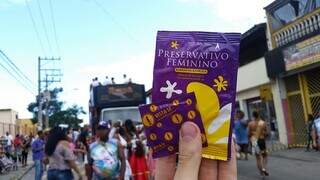 Pessoa segura preservativo masculino e feminino com bloco de Carnaval ao fundo (Foto: Divulgação)