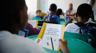 Criança em sala de aula segurando livro didático sob olhar da professora (Foto: Tânia Rego/Agência Brasil)
