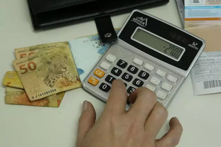 Mulher fazendo contas na calculadora, com dinheiro ao lado (Foto: Arquivo/Campo Grande News)