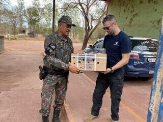 Policial militar e agente da Receita Federal segurando caixa com os pombos (Foto: Diário Corumbaense)