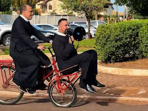 De bike e megafone, padres viram com&eacute;dia convidando para a novena